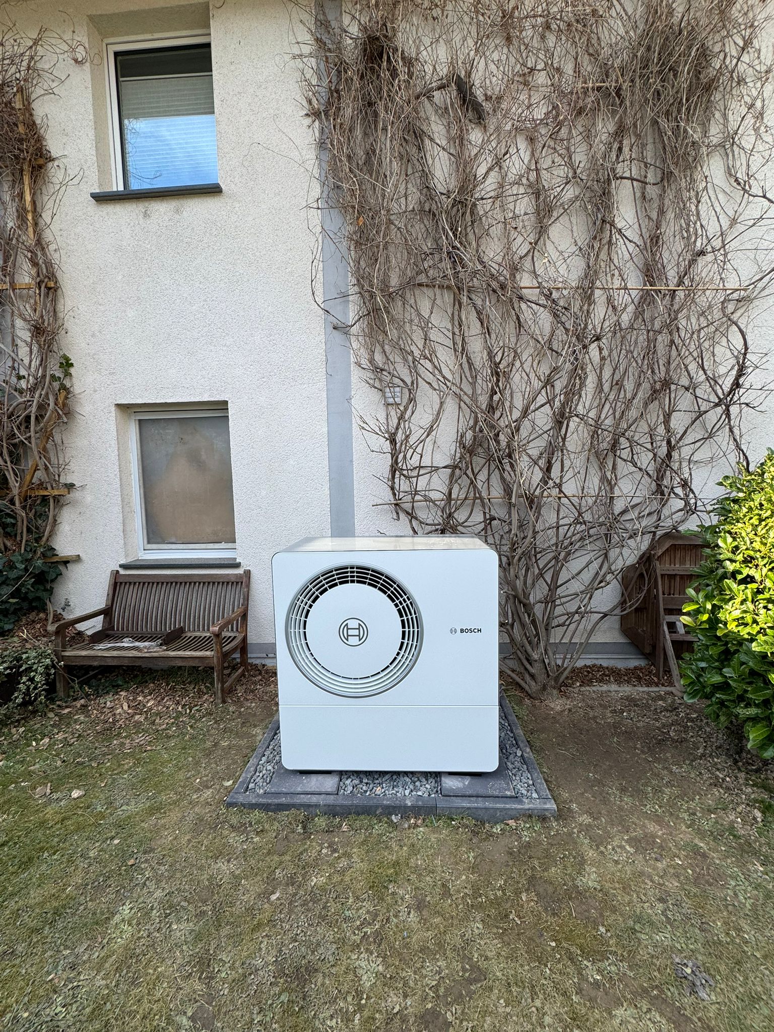 Eine Bosch-Wärmepumpe steht auf einem Fundament vor einer Hauswand mit Rankpflanzen, eingerahmt von zwei Holzbänken im Garten.